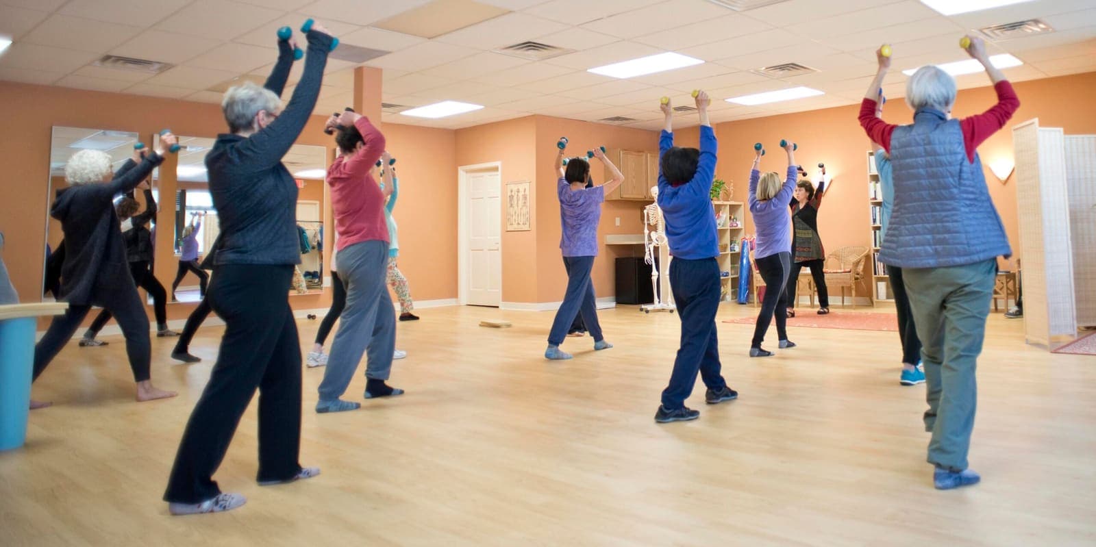A dozen students standing in a bright studio, reaching overhead with light hand weights